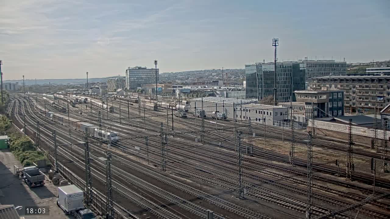 A vast railway yard with numerous train cars and tracks stretches out before a cityscape under a mostly clear sky.