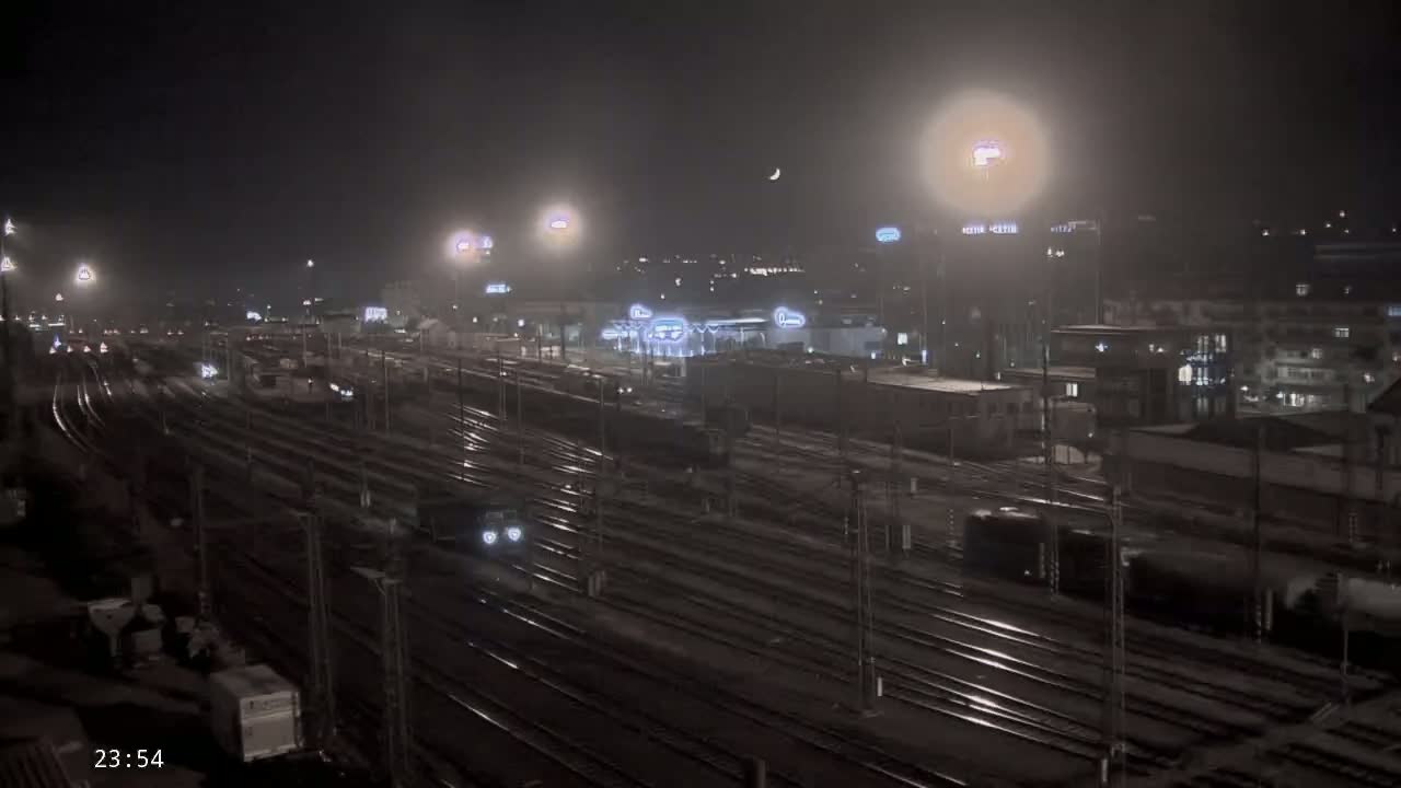 A nighttime view of a train yard with many tracks, some trains, and buildings in the background under hazy, possibly rainy conditions.