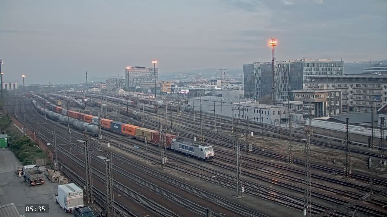 A freight train carrying orange and gray containers moves through a sprawling railway yard under an overcast sky.