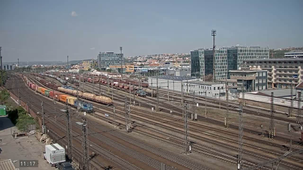 A sprawling railway yard filled with numerous freight trains under a clear, sunny sky, with modern buildings in the background.