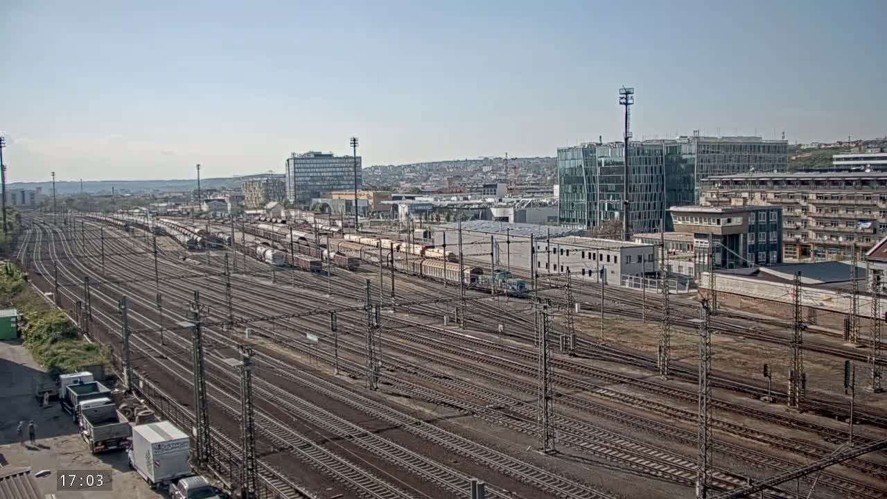 A sprawling railway yard with numerous tracks and freight cars under a clear, sunny sky, adjacent to several buildings.