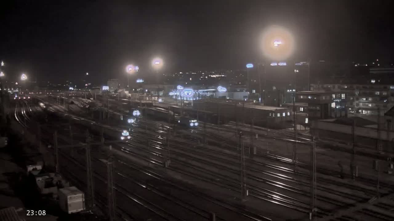 A nighttime view of a sprawling train yard with several trains and buildings in the background, lightly dusted with snow.