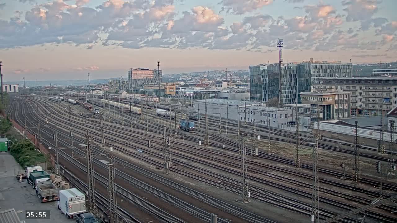 A sprawling railway yard with numerous tracks and parked freight cars is visible beneath a partly cloudy, dusky sky, with modern buildings in the background.