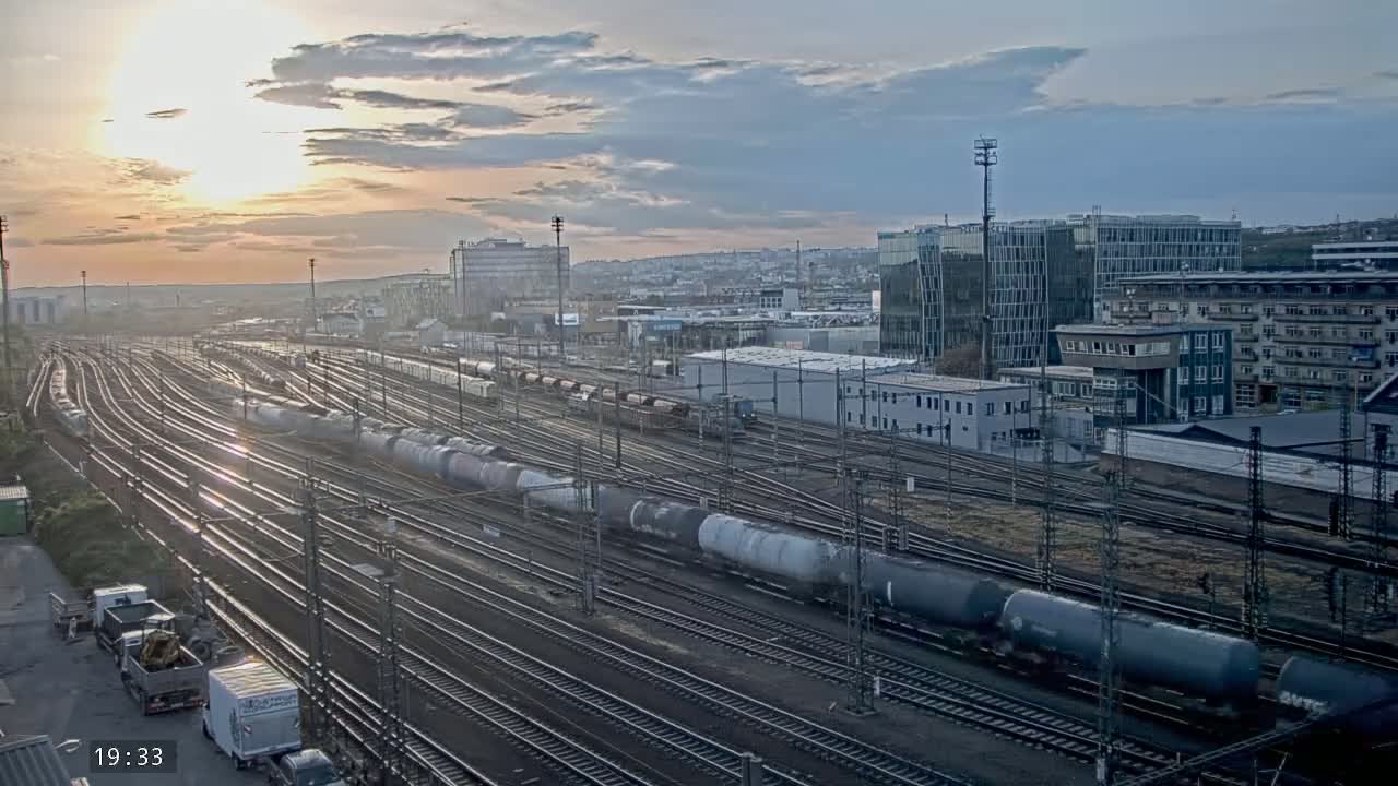 A sprawling rail yard filled with numerous freight cars and train tracks is seen at sunset under a partly cloudy sky.