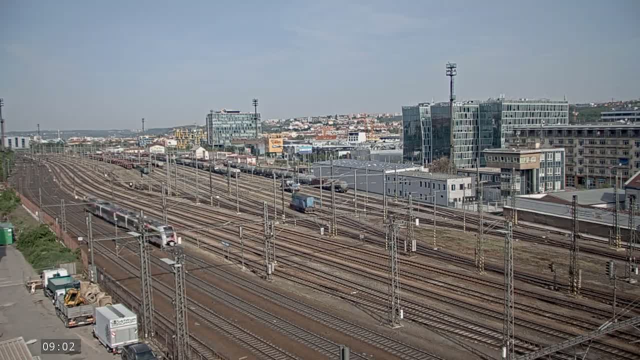 A sprawling railway yard with numerous tracks, trains, and modern buildings in the background under a clear, sunny sky.