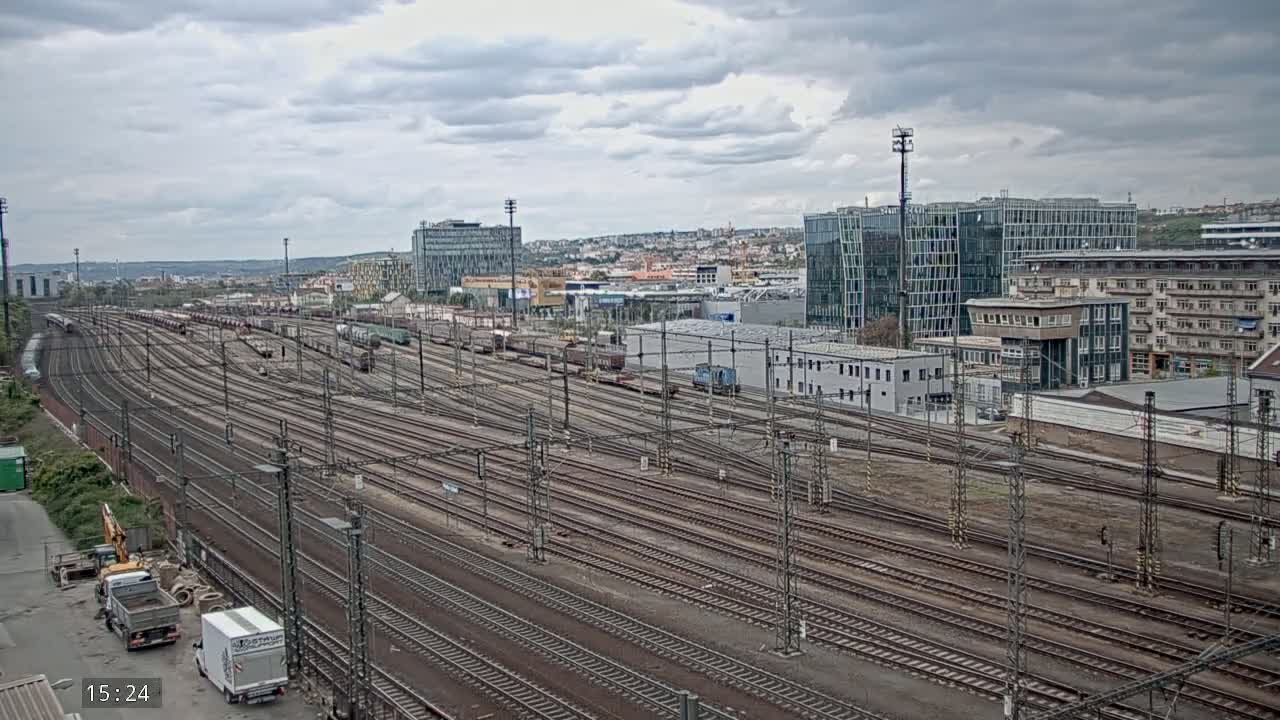 An extensive railway yard with numerous tracks and freight cars, under a cloudy sky, is situated near a city with modern and older buildings.