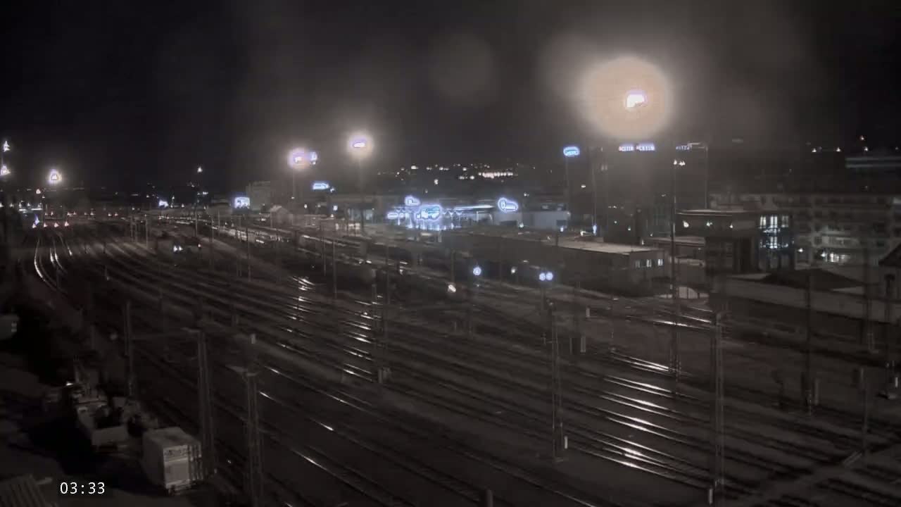 A nighttime view of a sprawling train yard with many tracks and some buildings in the background, under partly cloudy conditions.