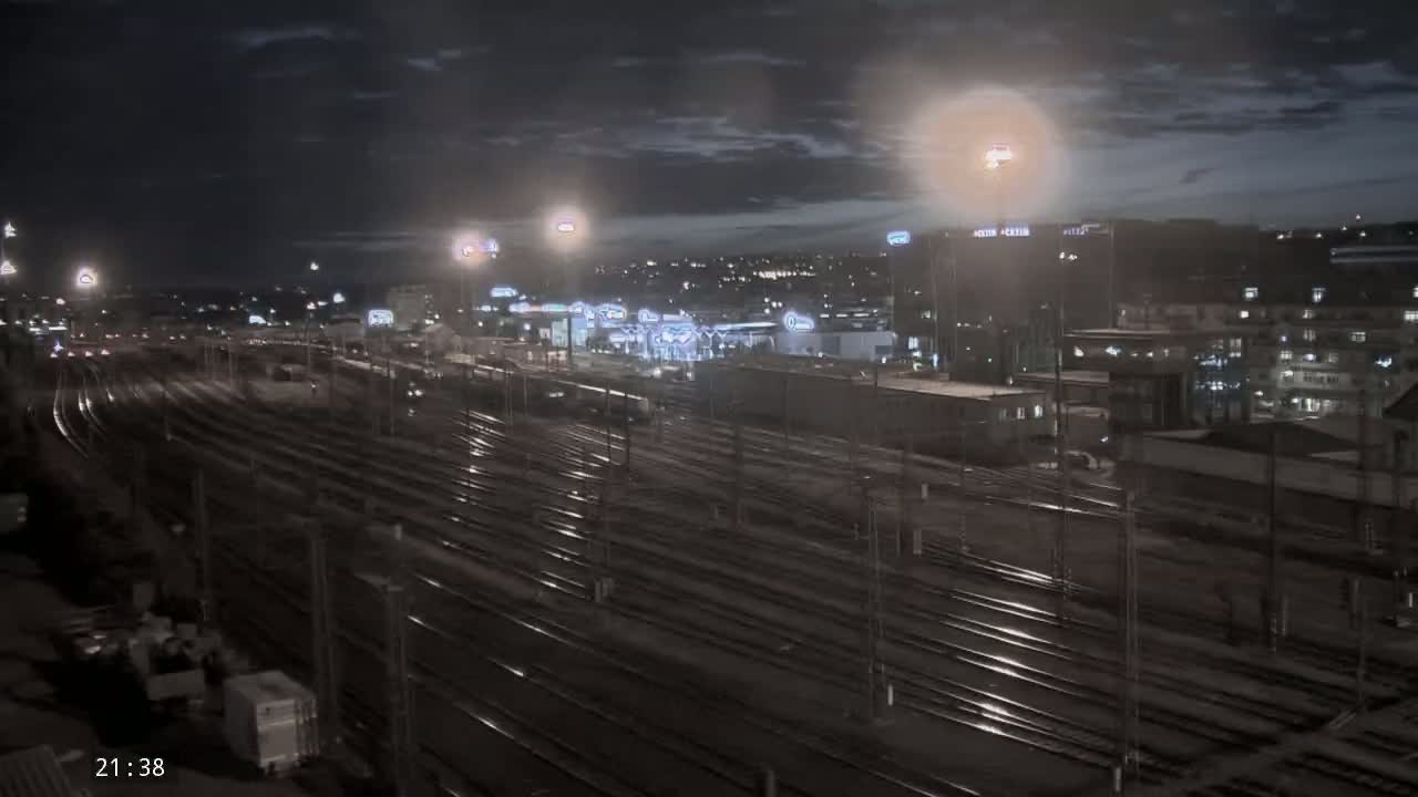 A nighttime view of numerous train tracks extending into the distance, with a city illuminated by lights in the background under a partly cloudy sky.