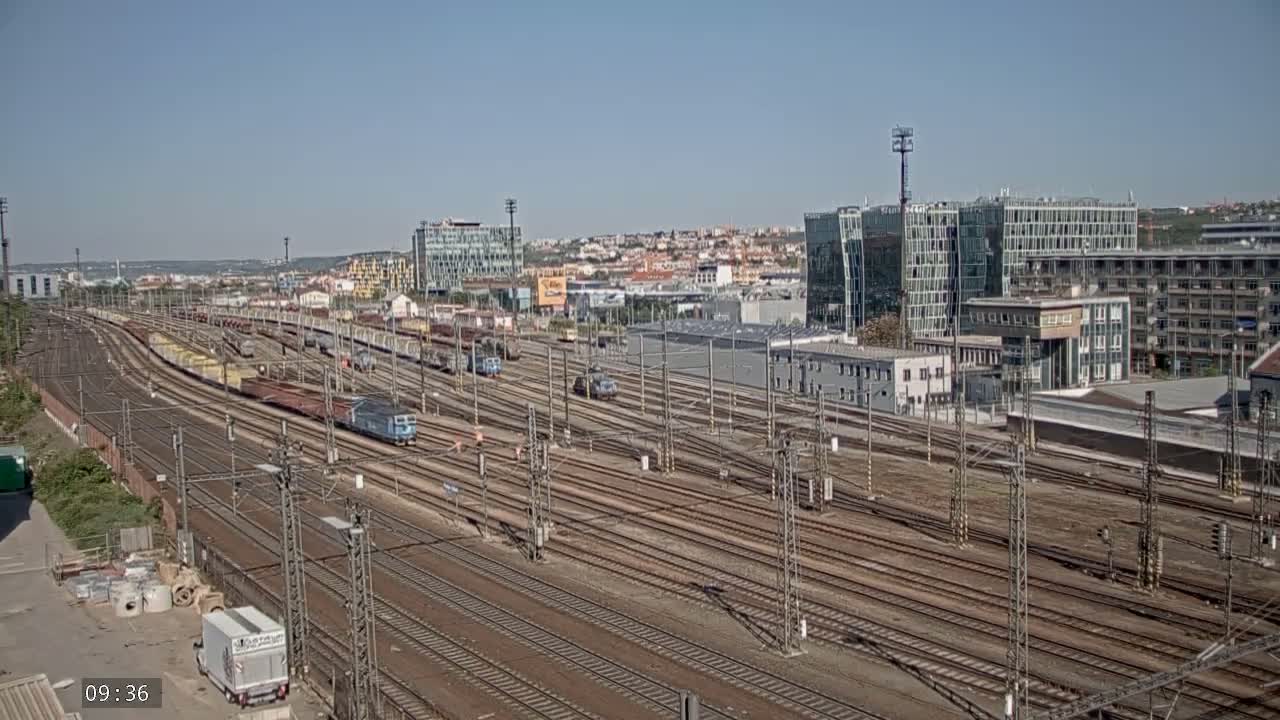 A sprawling railway yard with numerous train cars and tracks under a clear, sunny sky.