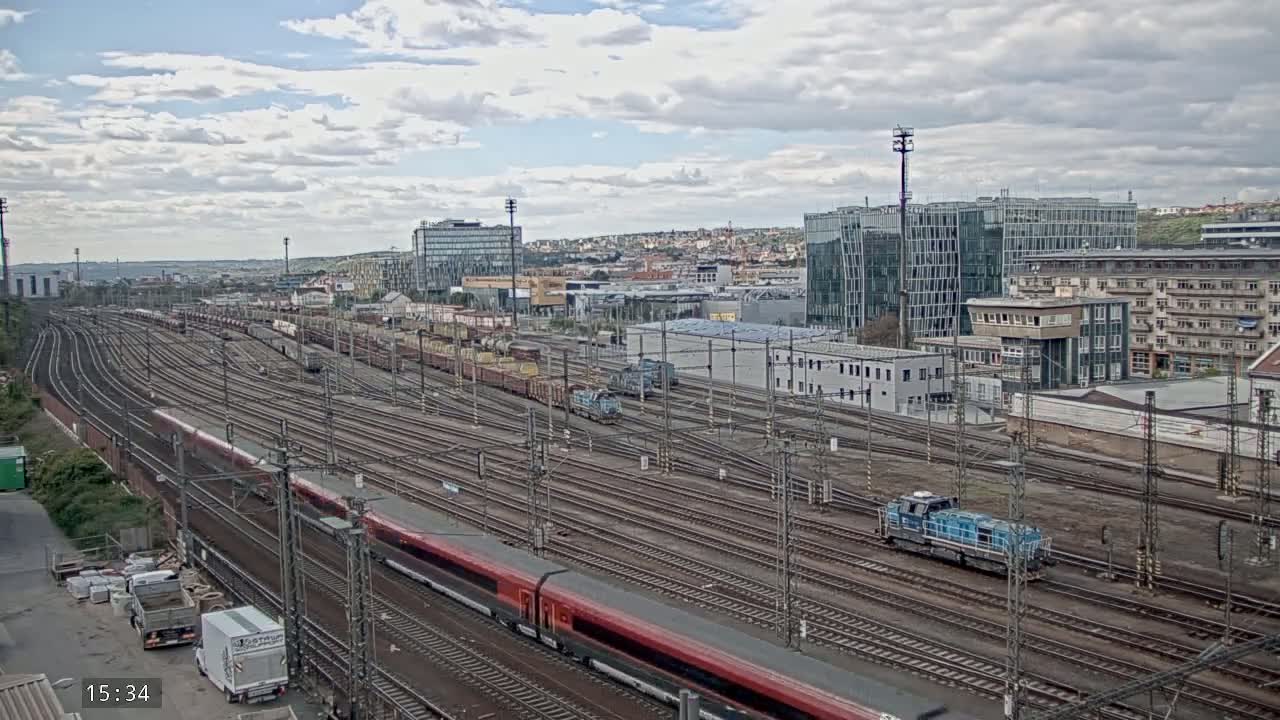 A large train yard with many tracks, several trains, and buildings in the background under a partly cloudy sky.