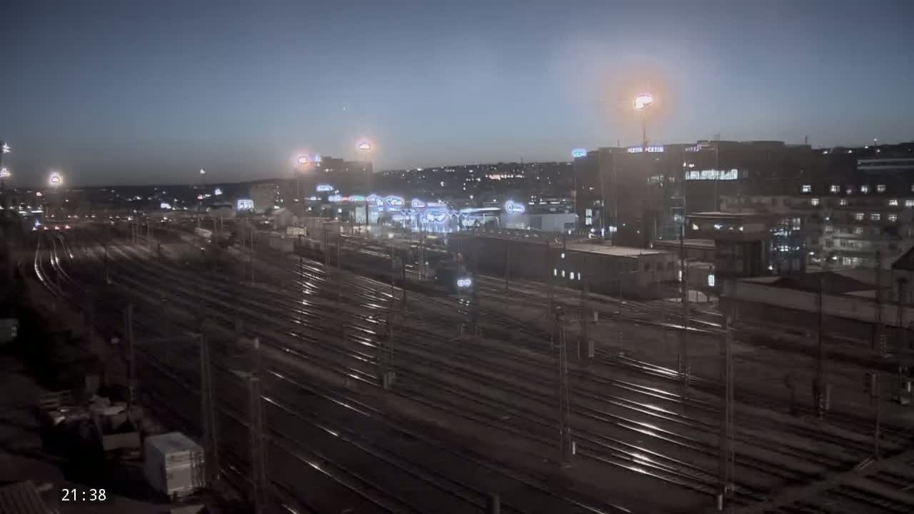 A nighttime, high-angle view of a train yard with many tracks and buildings in the background under a dark sky.