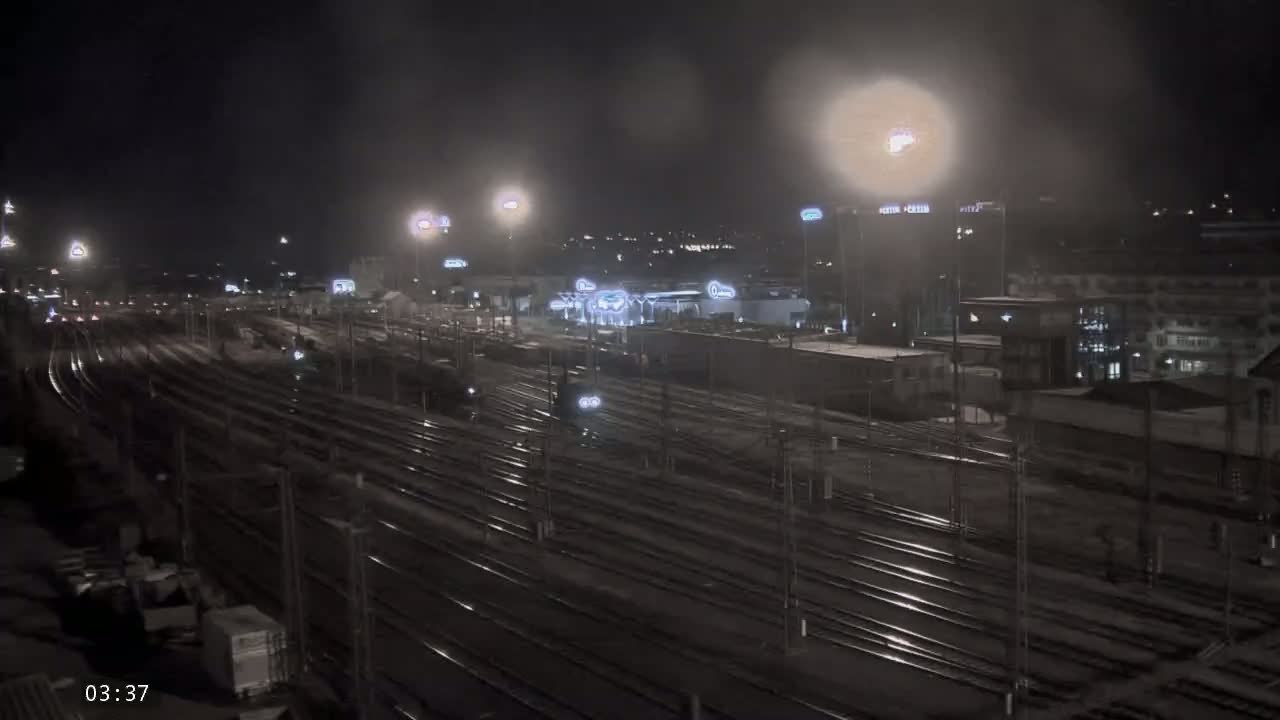 A nighttime view of a snow-covered train yard with several buildings in the background, lit by artificial lights.