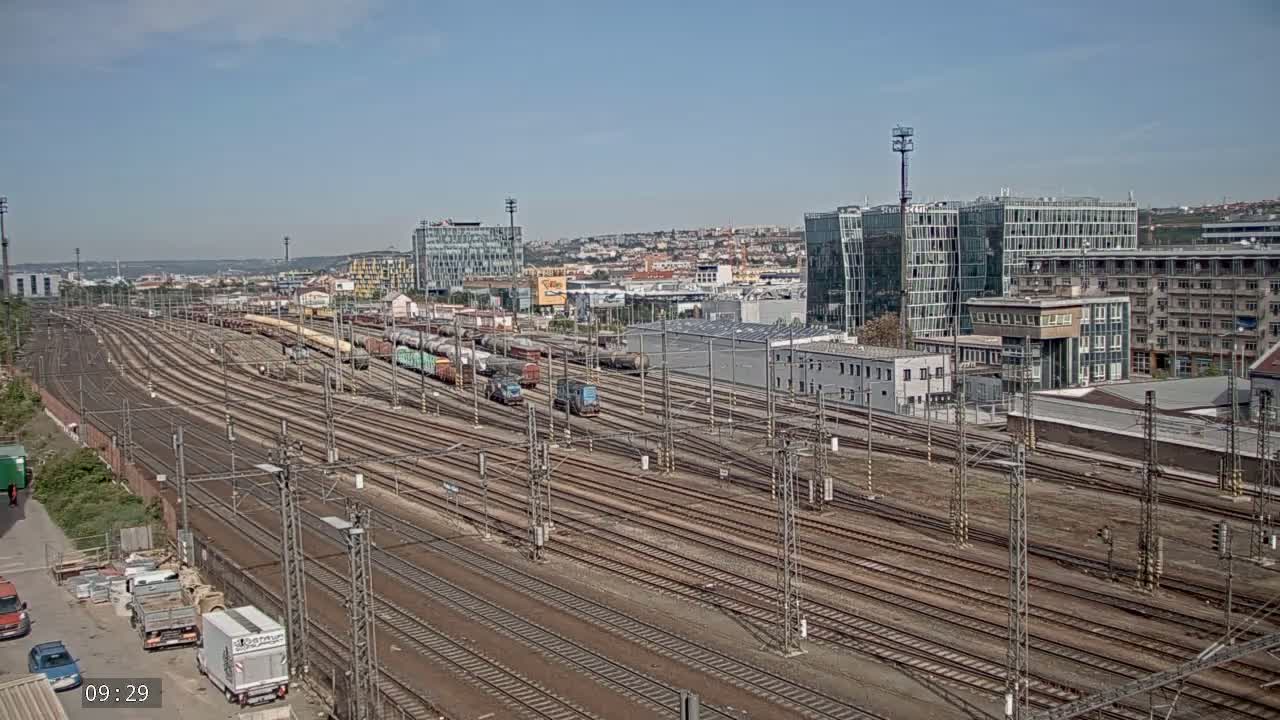 A wide view of a train yard with numerous railway tracks, freight trains, and buildings under a clear sky.
