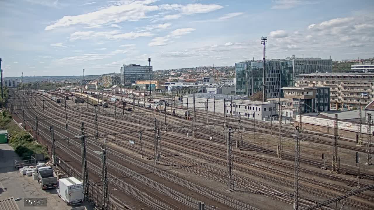 A sprawling railway yard with numerous tracks and parked freight cars stretches towards a cityscape under a partly cloudy sky.