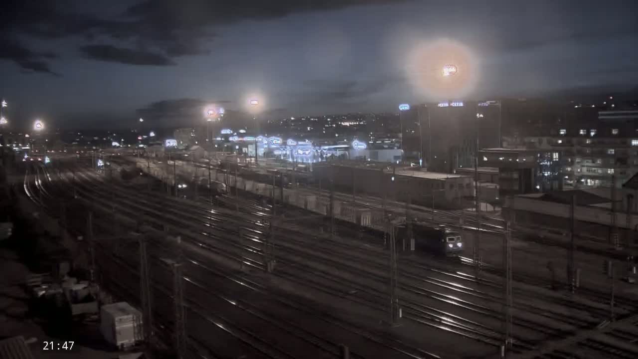 A nighttime view of a train station with many railway tracks, a few trains, and city buildings in the background under a cloudy sky.