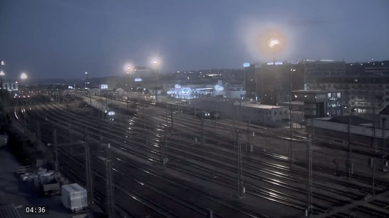 A nighttime view of a large train yard with numerous tracks extending into the distance, surrounded by city buildings under a dimly lit sky.