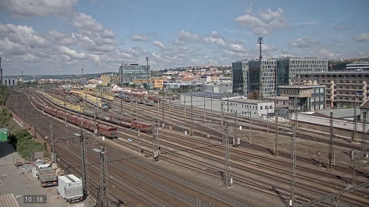 Numerous freight trains are parked on a large rail yard under a partly cloudy sky, with modern buildings visible in the background.