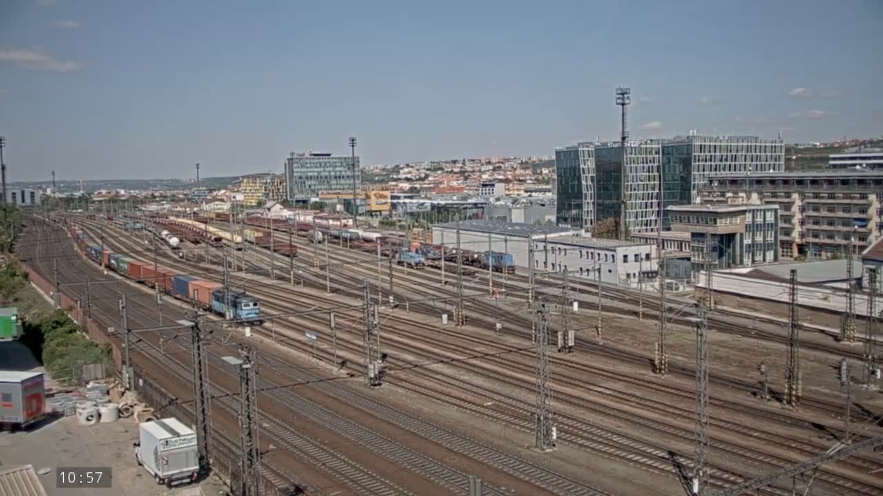 A sprawling rail yard filled with numerous train cars and locomotives under a clear, sunny sky, adjacent to a city with modern and older buildings.