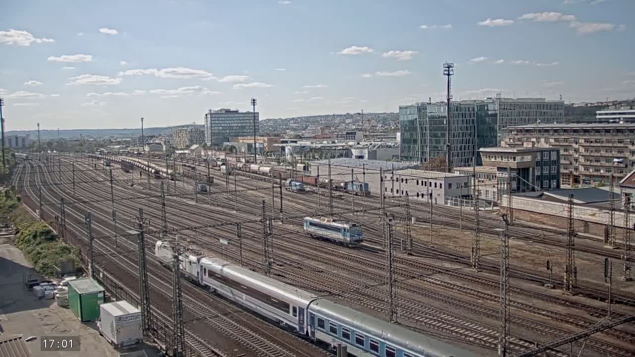 A long expanse of train tracks in a railway yard, with several trains and buildings visible in the background under a partly cloudy sky.
