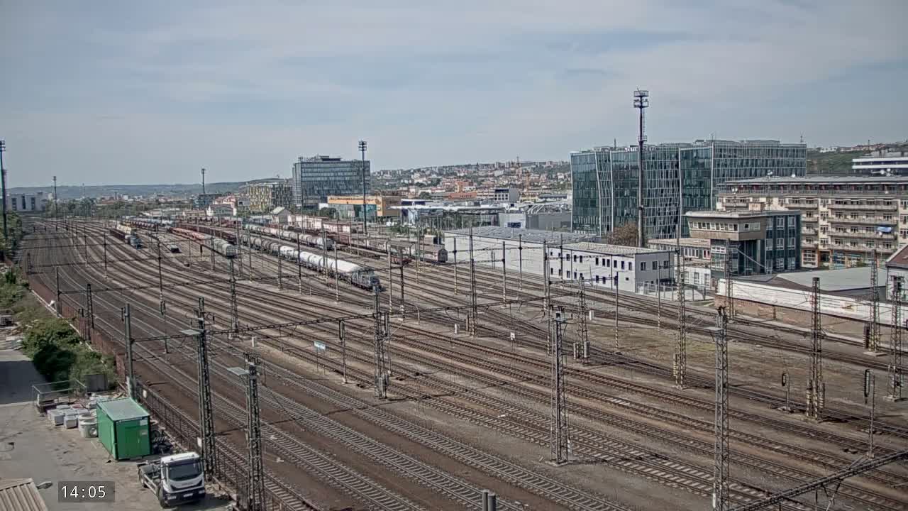 A sprawling railway yard with numerous train cars and tracks extends towards a cityscape under a partly cloudy sky.