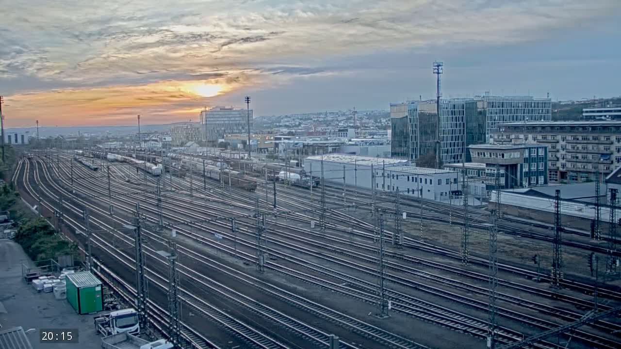 A vast network of train tracks stretches out beneath a sunset sky, with numerous freight cars parked alongside buildings in the background.