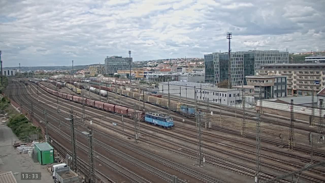 A light-blue locomotive leads a long freight train through a sprawling railway yard under a cloudy sky.