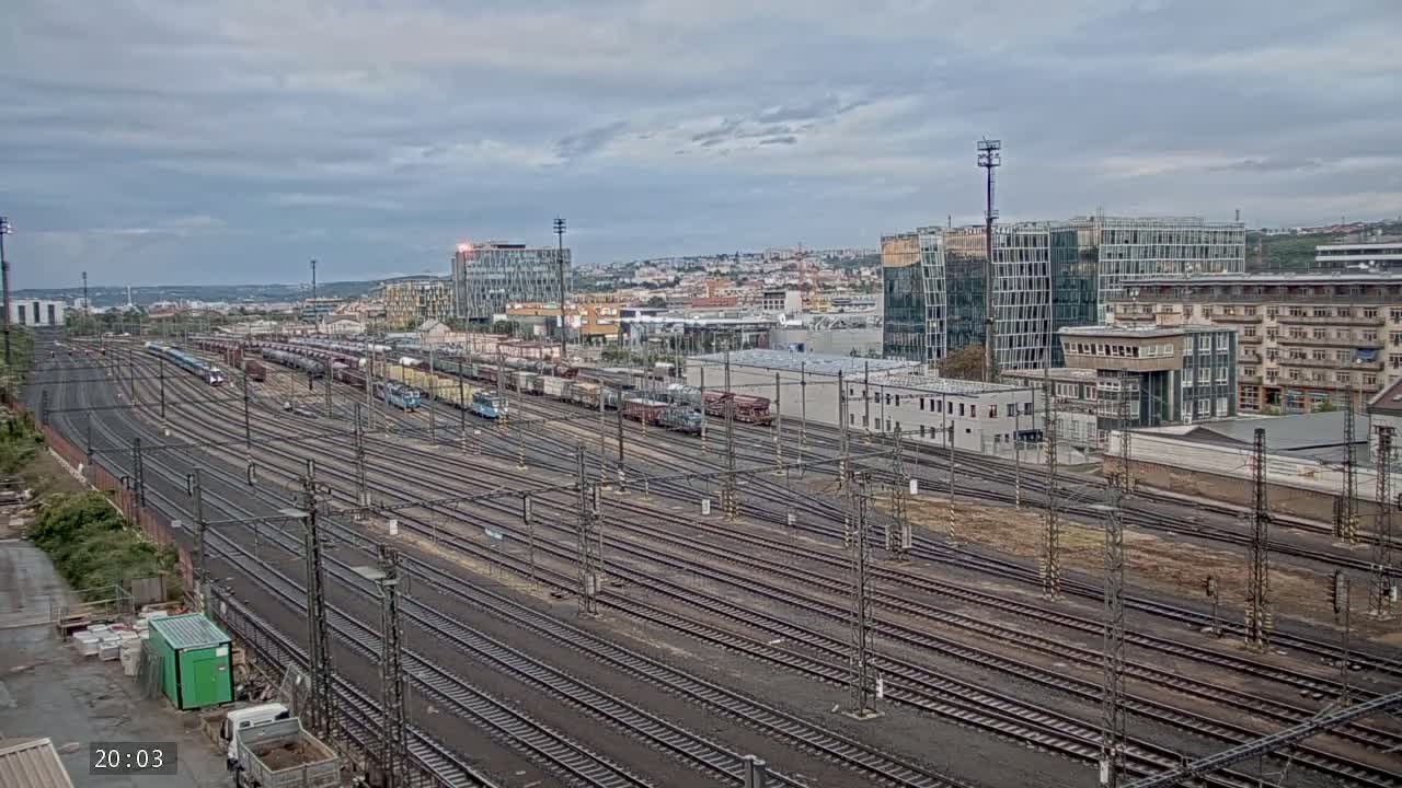 Numerous train tracks extend into the distance under an overcast sky, with a collection of trains and cargo containers parked nearby and buildings in the background.