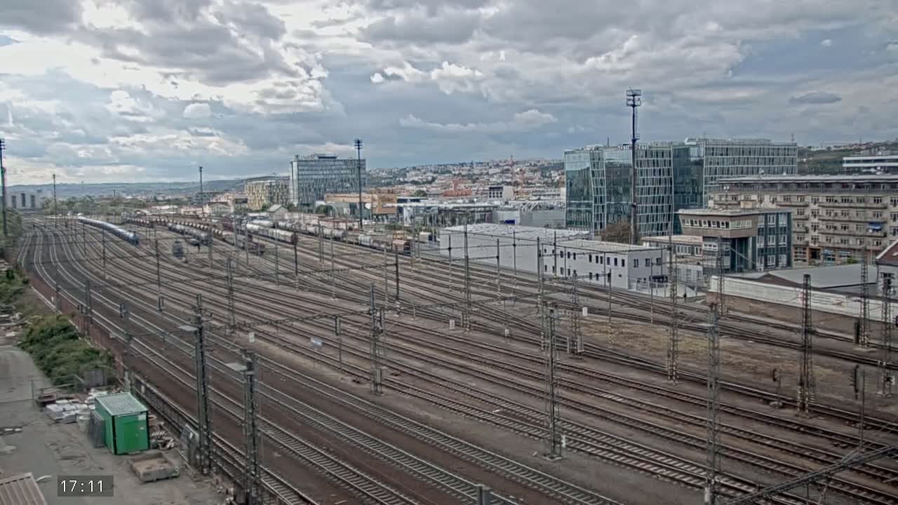 A large railway yard with many train tracks extends into the distance under a cloudy sky, with modern buildings visible in the background.
