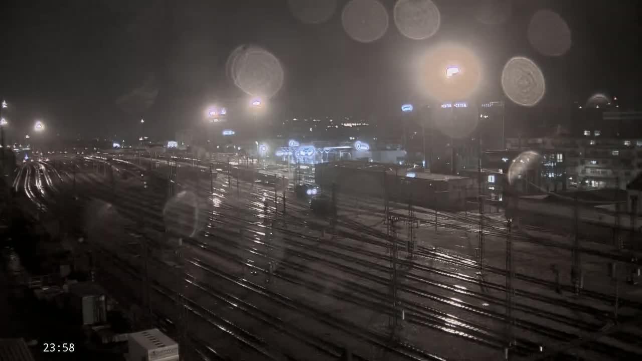 A nighttime view of a rain-slicked railway yard with numerous tracks and buildings in the background under a dark, rainy sky.