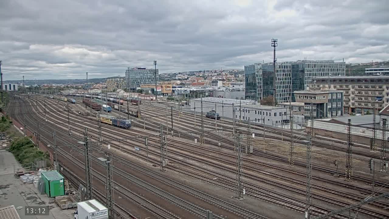 A large railway yard with numerous train tracks and several trains, under a cloudy sky, is situated next to a city with modern and older buildings.