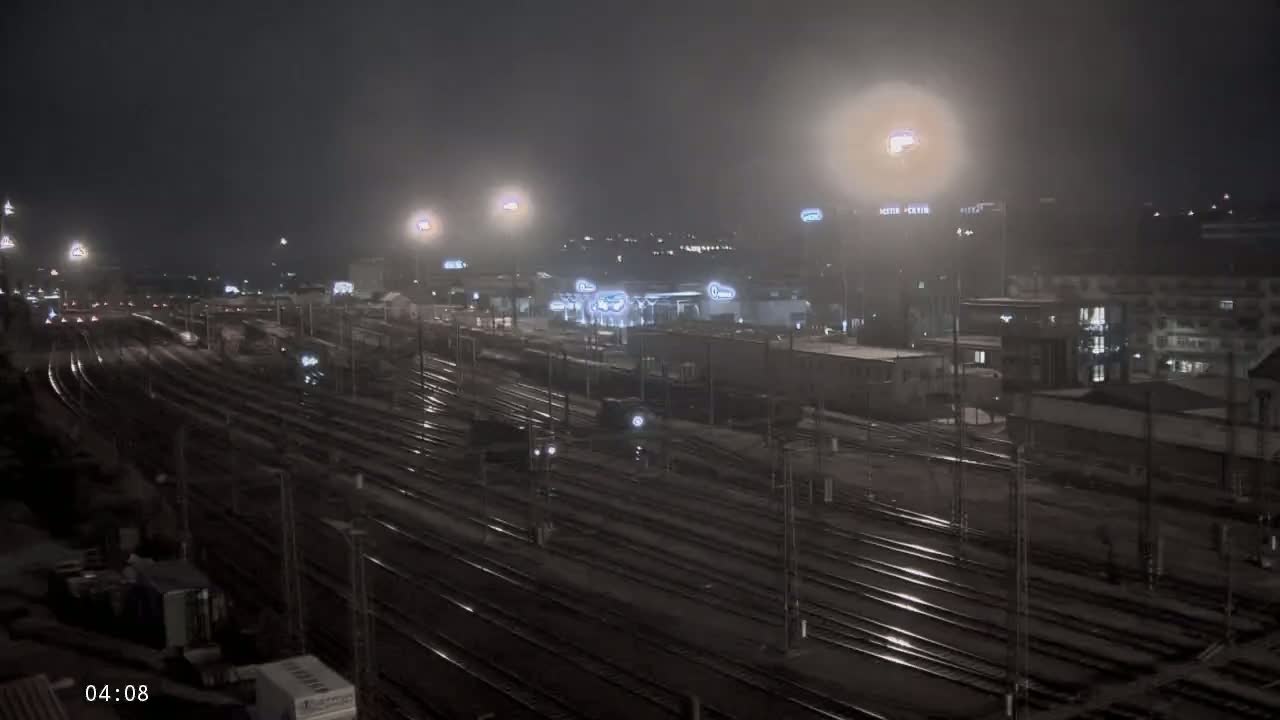 A nighttime view of a sprawling, wet train yard with buildings and lights in the background under a hazy sky.