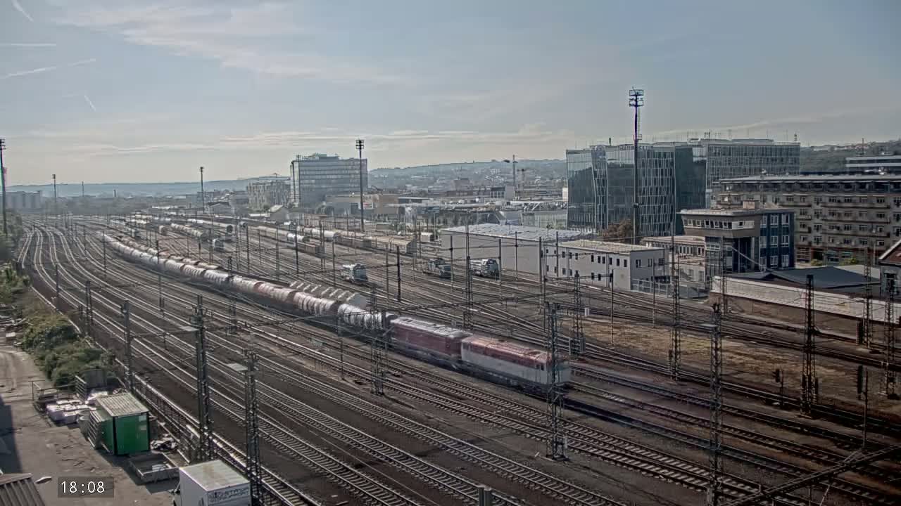A long freight train travels along a vast network of train tracks under a partly sunny sky, with numerous buildings and structures visible in the background.