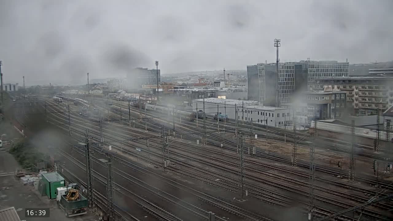 A rainy view of a large railway yard with numerous tracks, surrounded by buildings in a city.