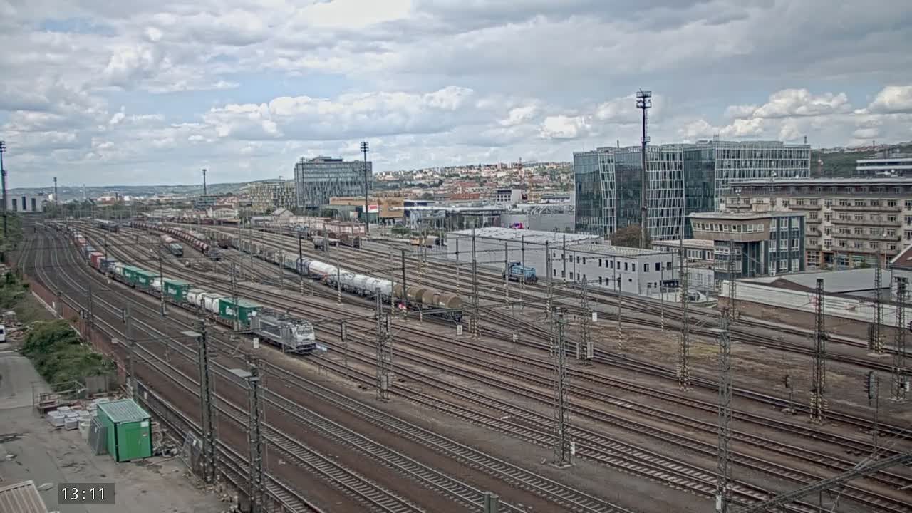 A large railway yard with numerous train cars and tracks, under a partly cloudy sky, is situated near several modern buildings.