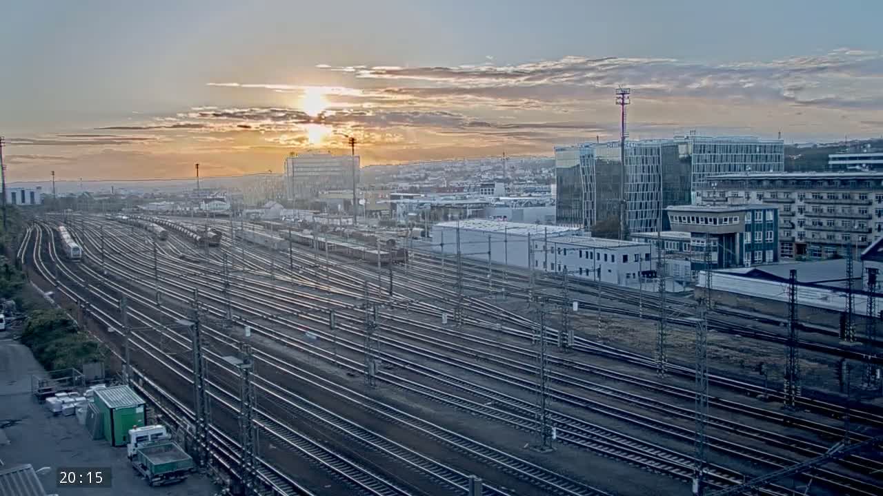 Many train tracks extend into the distance beneath a setting sun, surrounded by industrial buildings and a city skyline.