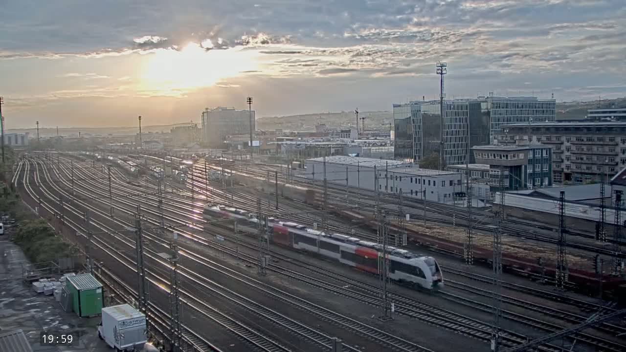 A long passenger train travels across a sprawling railway yard at sunset under a partly cloudy sky.