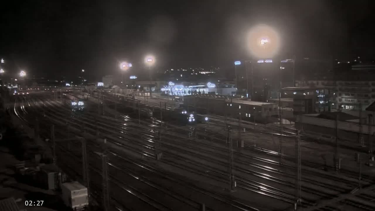 A nighttime, dimly lit view of a large train yard with many tracks and trains, under slightly hazy conditions.