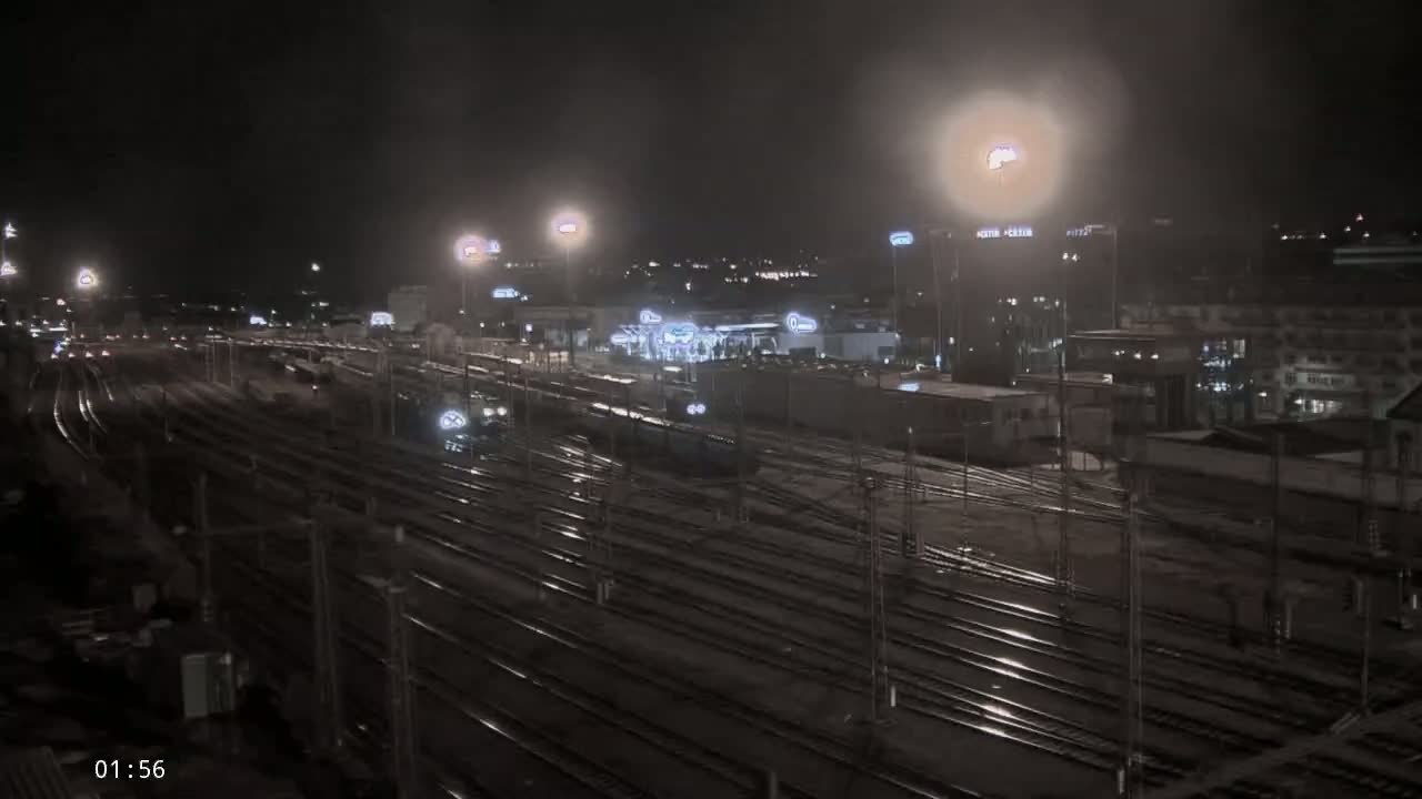 A nighttime view of a rain-slicked railway yard with several trains and buildings illuminated by artificial lights in the background.