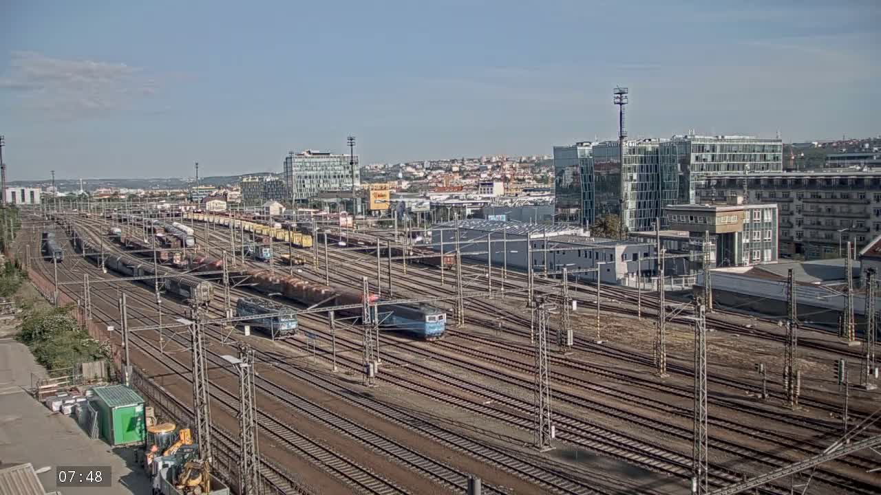 A sprawling railway yard with numerous freight trains and locomotives under a clear, sunny sky, situated near a cityscape.