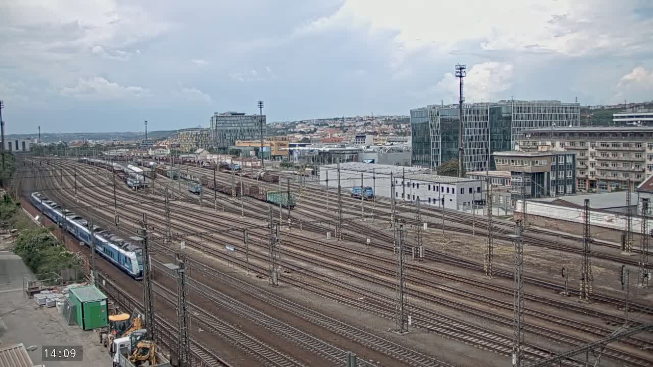 A long train travels past numerous railway tracks and freight cars under a cloudy sky, with buildings and a cityscape visible in the background.