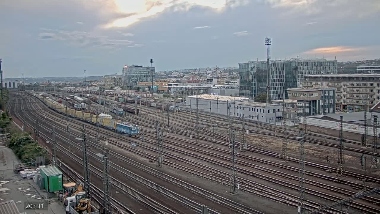 A sprawling railway yard with numerous train cars and tracks under an overcast sky, situated near a city with modern and older buildings.