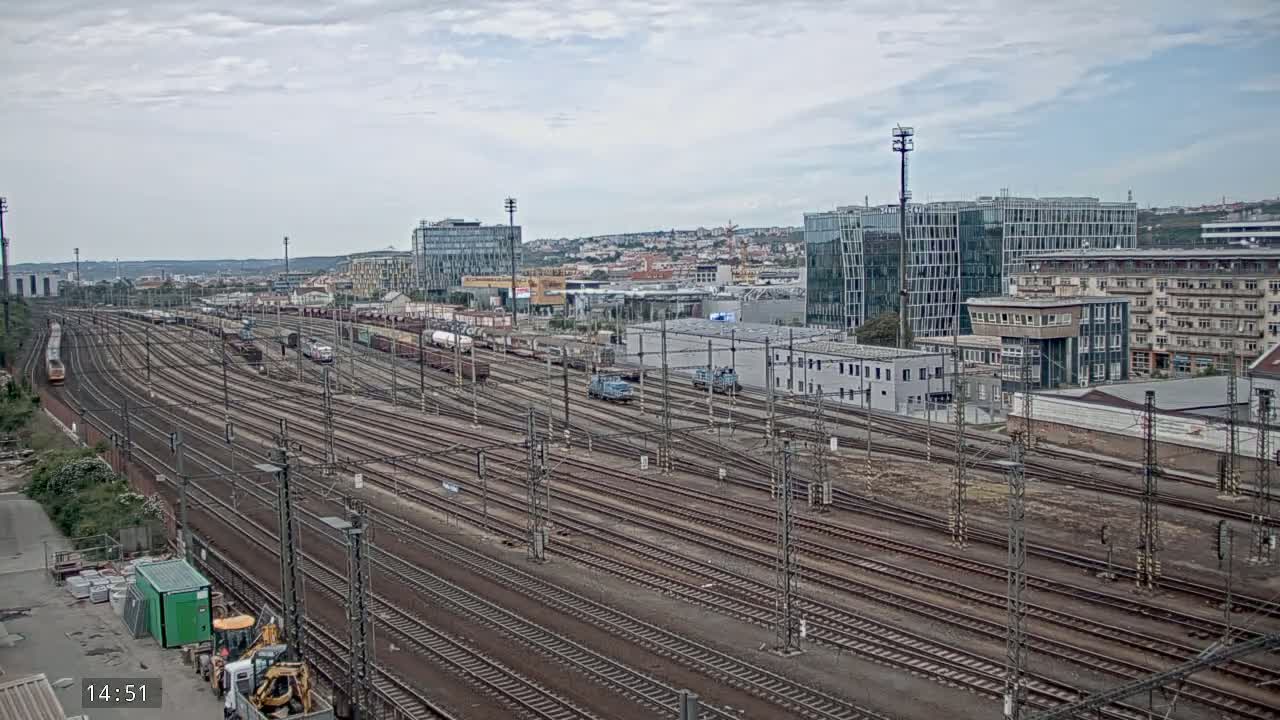 A sprawling railway yard with numerous tracks and parked freight cars is seen under an overcast sky, adjacent to modern and older buildings.