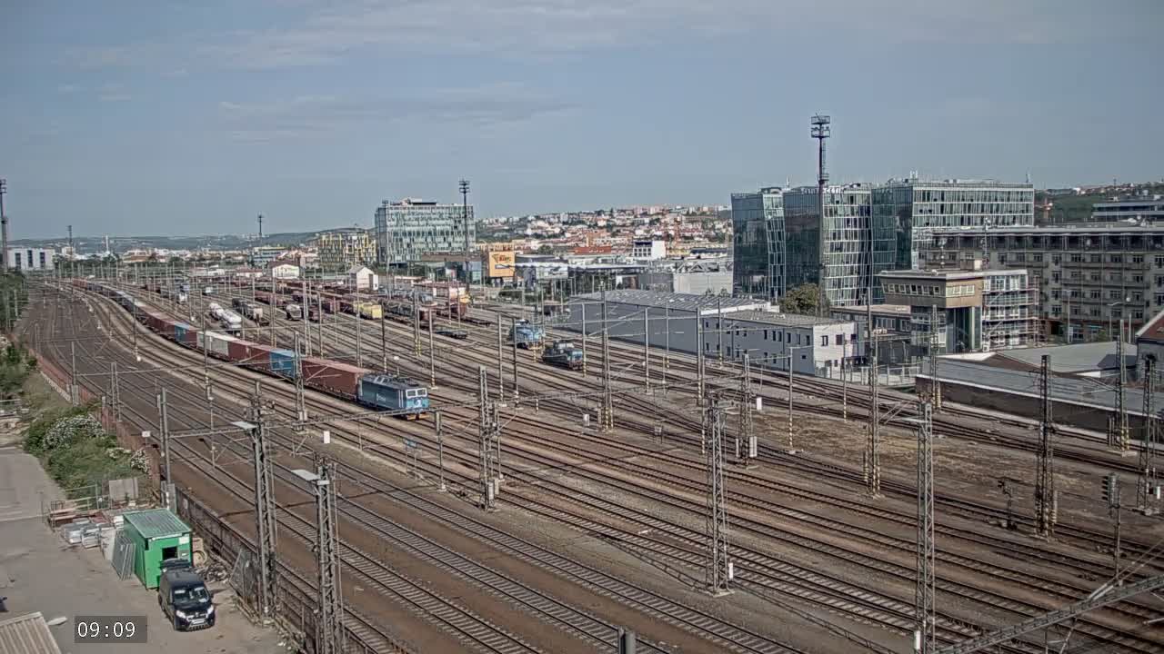 A sprawling railway yard with numerous train cars and locomotives, situated near a city skyline under a mostly sunny sky.