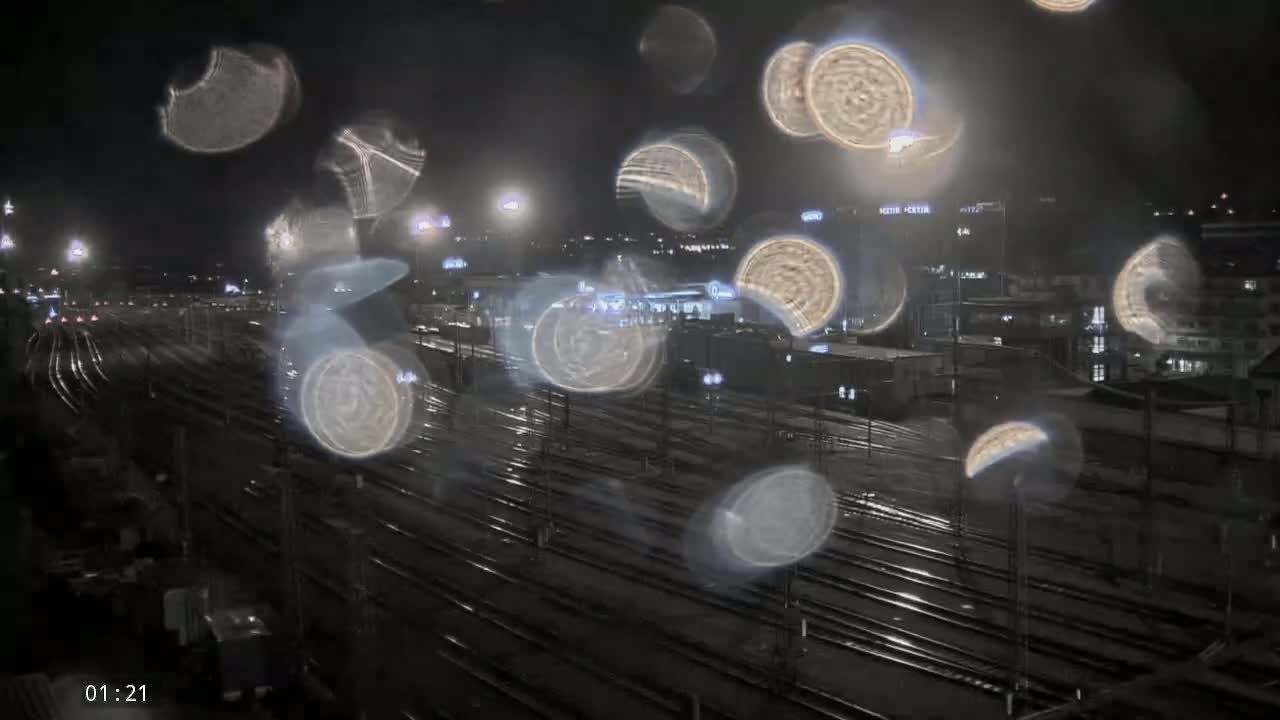 Rain-streaked glass obscures a nighttime view of a sprawling railway yard illuminated by numerous lights.