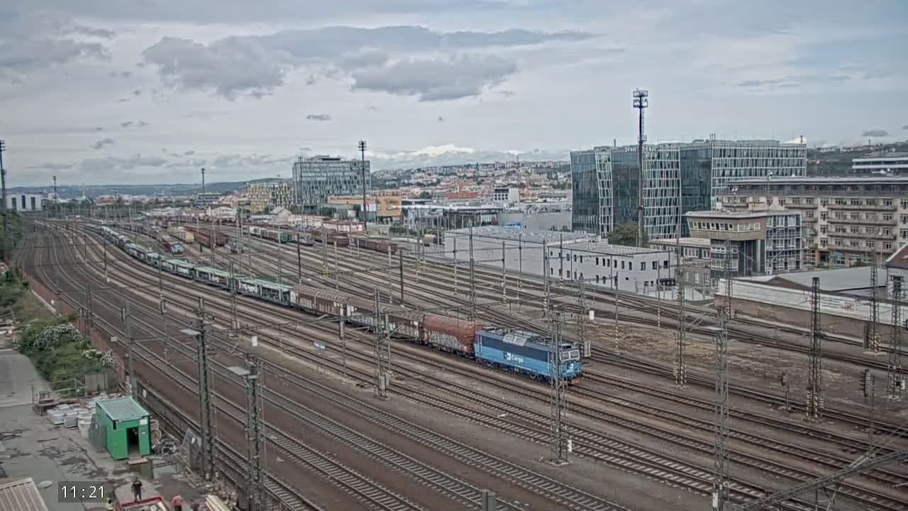 A long freight train is visible on a sprawling railway network under a cloudy sky, near a cityscape of modern and older buildings.