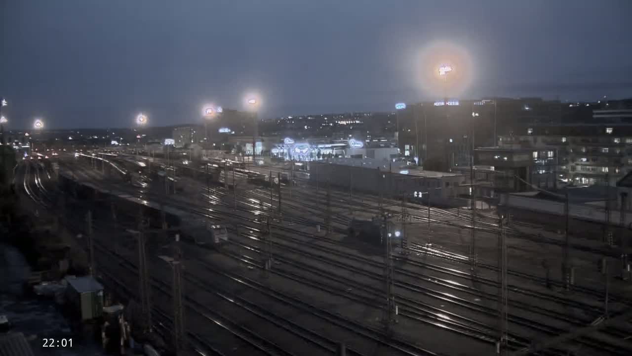 A nighttime, rain-slicked railway yard with numerous tracks extends toward a city skyline illuminated by streetlights.
