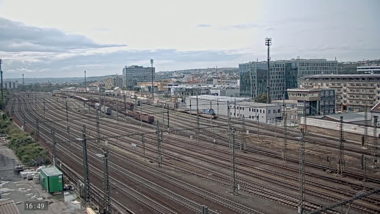 An expansive railway yard with numerous train tracks stretches out before a cityscape under a mostly cloudy sky.