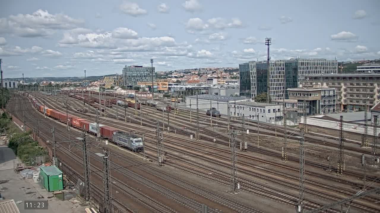 A large freight train moves along numerous railway tracks under a partly cloudy sky, passing by a complex of buildings.