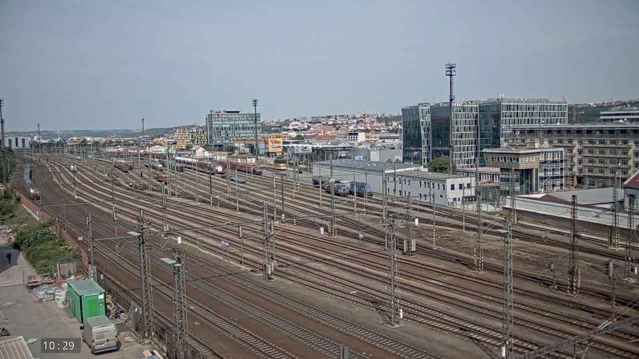 A wide shot on a sunny day shows numerous railway tracks extending into the distance, past a collection of freight cars and a cityscape with modern and older buildings.