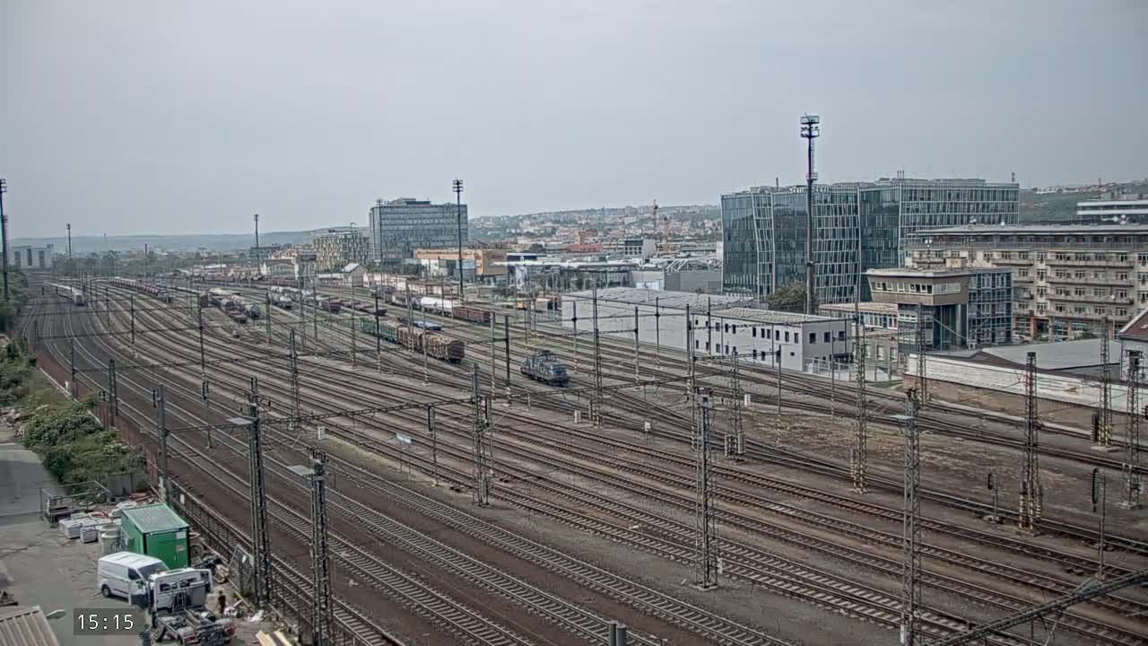 A sprawling railway yard with numerous train tracks and freight cars, under an overcast sky, is situated near a cityscape of modern and older buildings.